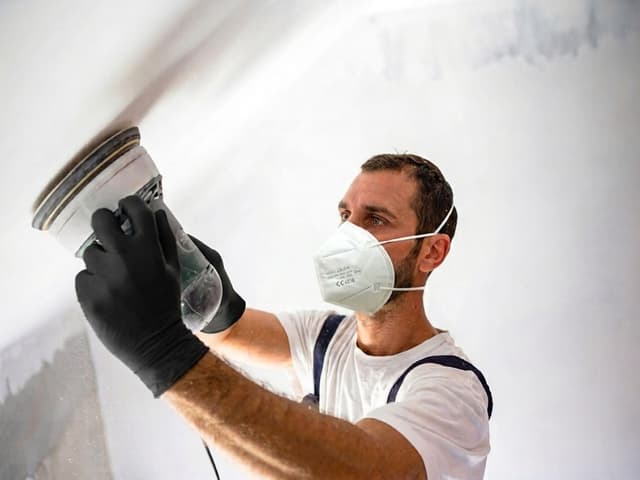 A tradesperson wearing a protective mask and gloves while using a powered sanding tool
